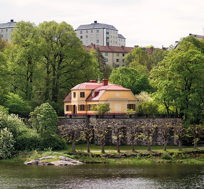 En gul 1700-talsbyggnad med rött tak vid vattnet, omgivet av grönska. I bakgrunden skymtar moderna höghus.