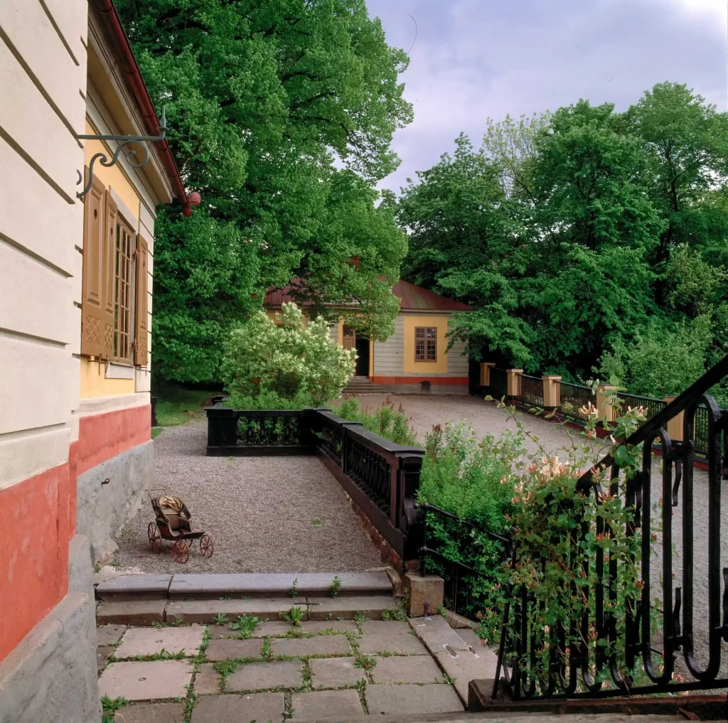 Från trappan ned från en byggnad syns ett separat hus en bit bort mot trädgården, en grusad gårdsplan emellan.